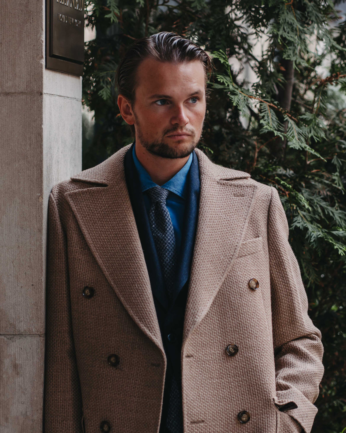 A man wearing a beige double-breasted overcoat over a blue shirt and patterned tie, standing outdoors next to a stone wall and green foliage.