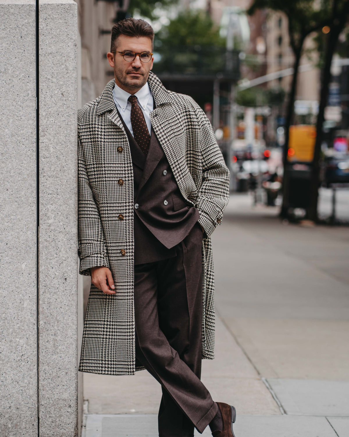 A man wearing a stylish brown and gray plaid single-breasted overcoat, matching brown suit, white shirt, and a brown patterned tie, standing on a city sidewalk next to a stone wall.