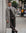 A man wearing a stylish brown and gray plaid single-breasted overcoat, matching brown suit, white shirt, and a brown patterned tie, standing on a city sidewalk next to a stone wall.