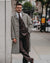 A man wearing a stylish brown and gray plaid single-breasted overcoat, matching brown suit, white shirt, and a brown patterned tie, standing on a city sidewalk next to a stone wall.