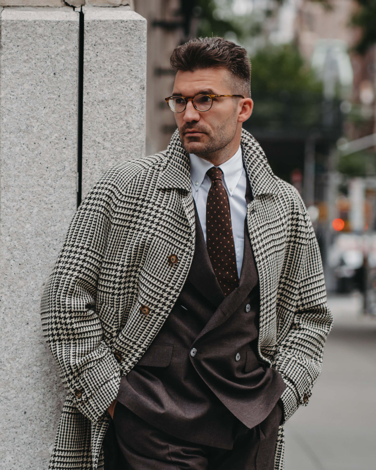 A man wearing a classic black and white houndstooth overcoat, a dark brown double-breasted suit, a white dress shirt, and a brown patterned tie, standing outdoors next to a gray stone wall.