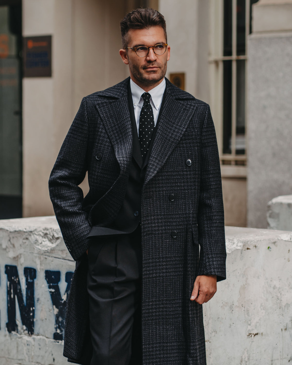 A man wearing a dark charcoal gray double-breasted plaid overcoat, black suit, a white dress shirt, and a black patterned tie, standing outdoors in front of a stone building.