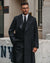 A man wearing a dark charcoal gray double-breasted plaid overcoat, black suit, a white dress shirt, and a black patterned tie, standing outdoors in front of a stone building.