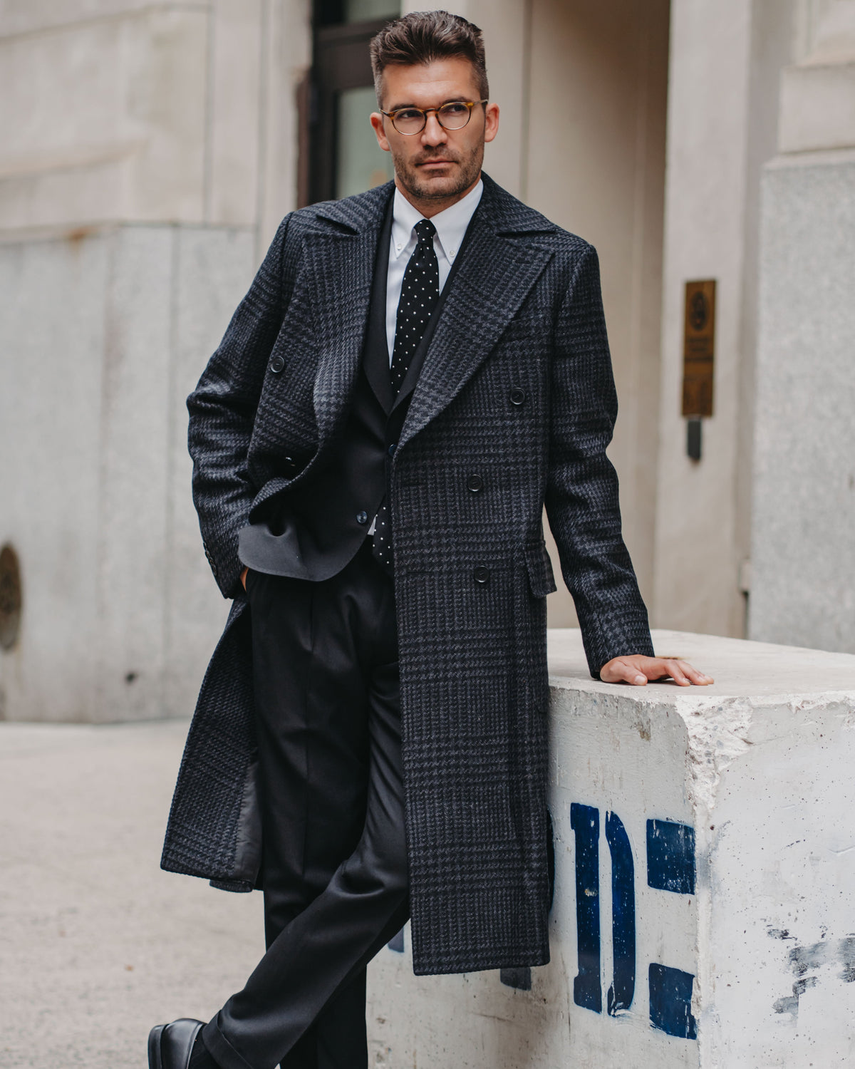 A man wearing a dark charcoal gray double-breasted plaid overcoat, black suit, white shirt, and patterned tie leans against a concrete barrier outdoors.