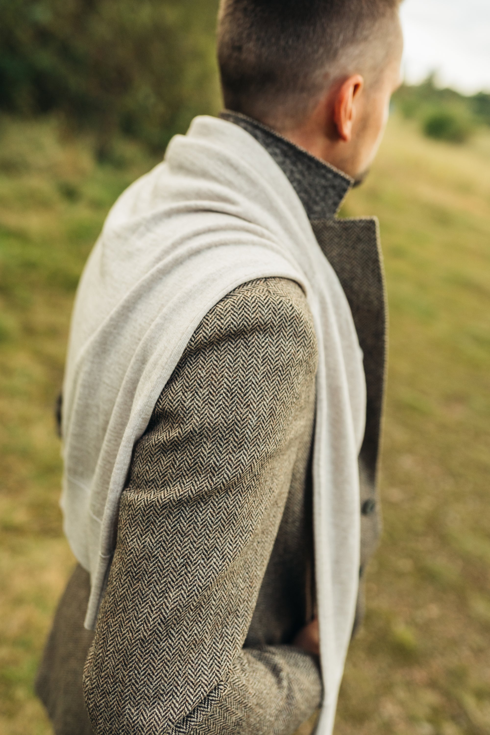 Person wearing a brown herringbone tweed jacket and beige sweater over the shoulders in a natural setting