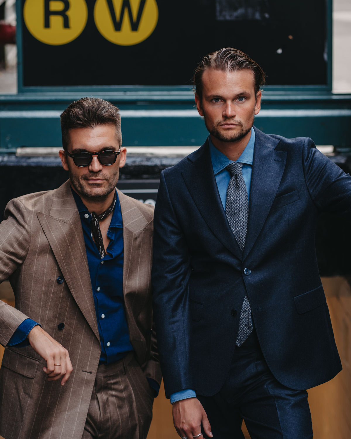 Two men posing in front of a New York City subway sign. The man on the right wears a dark navy blue suit with a light blue dress shirt and a patterned tie.