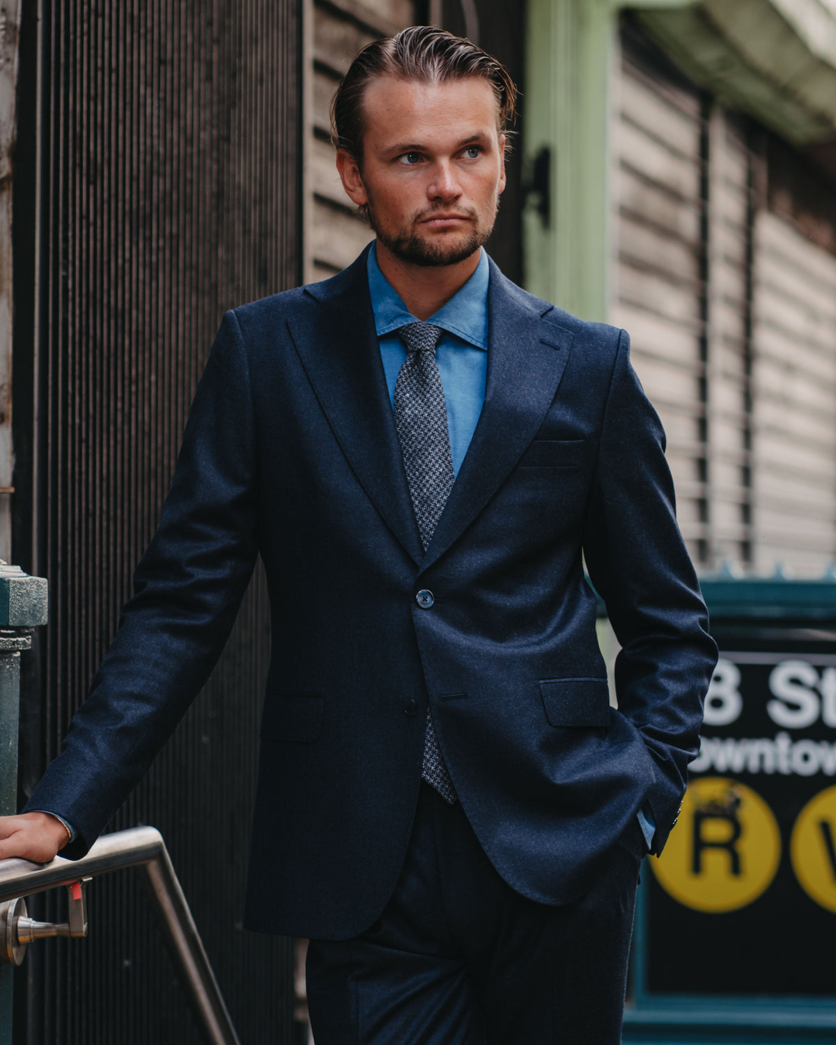 A man wearing a dark navy blue suit, a light blue dress shirt, and a patterned tie, standing in an urban outdoor setting.