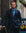 A man wearing a dark navy blue suit, a light blue dress shirt, and a patterned tie, standing in an urban outdoor setting.