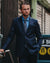 A man wearing a dark navy blue suit, a light blue dress shirt, and a patterned tie, standing in an urban outdoor setting.