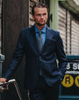 A man wearing a dark navy blue suit, a light blue dress shirt, and a patterned tie, standing in an urban outdoor setting.