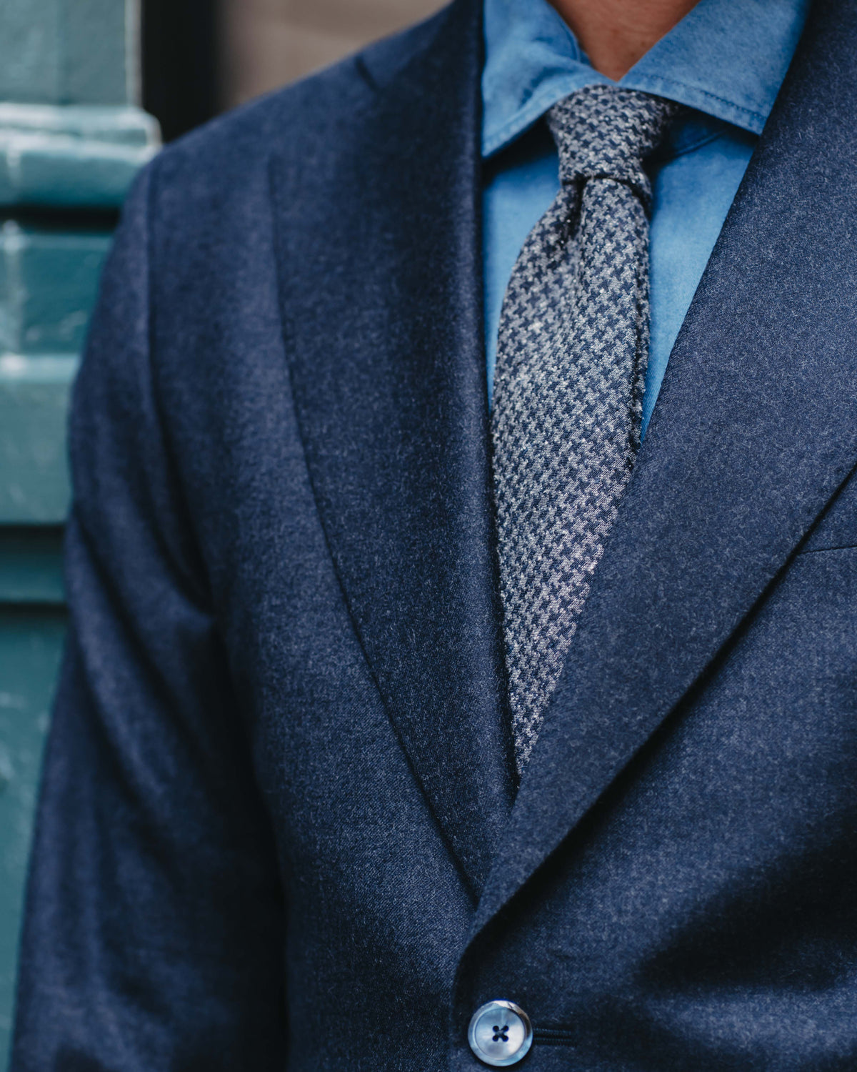 A close-up of a man wearing a textured navy blue suit jacket, a light blue dress shirt, and a patterned gray and white tie.