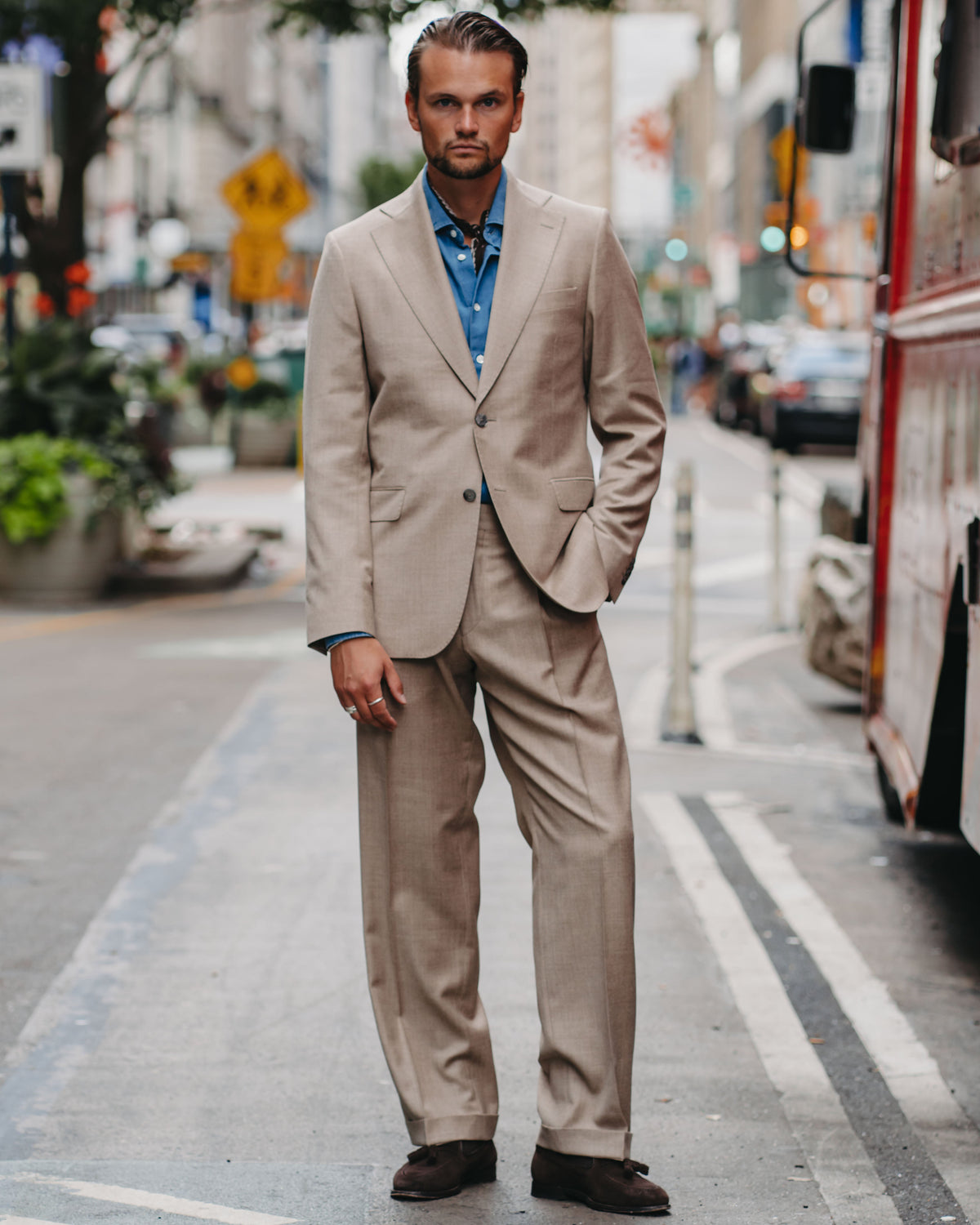 A man wearing a beige two-piece suit with a light blue button-down shirt and dark brown loafers, standing on a city street.