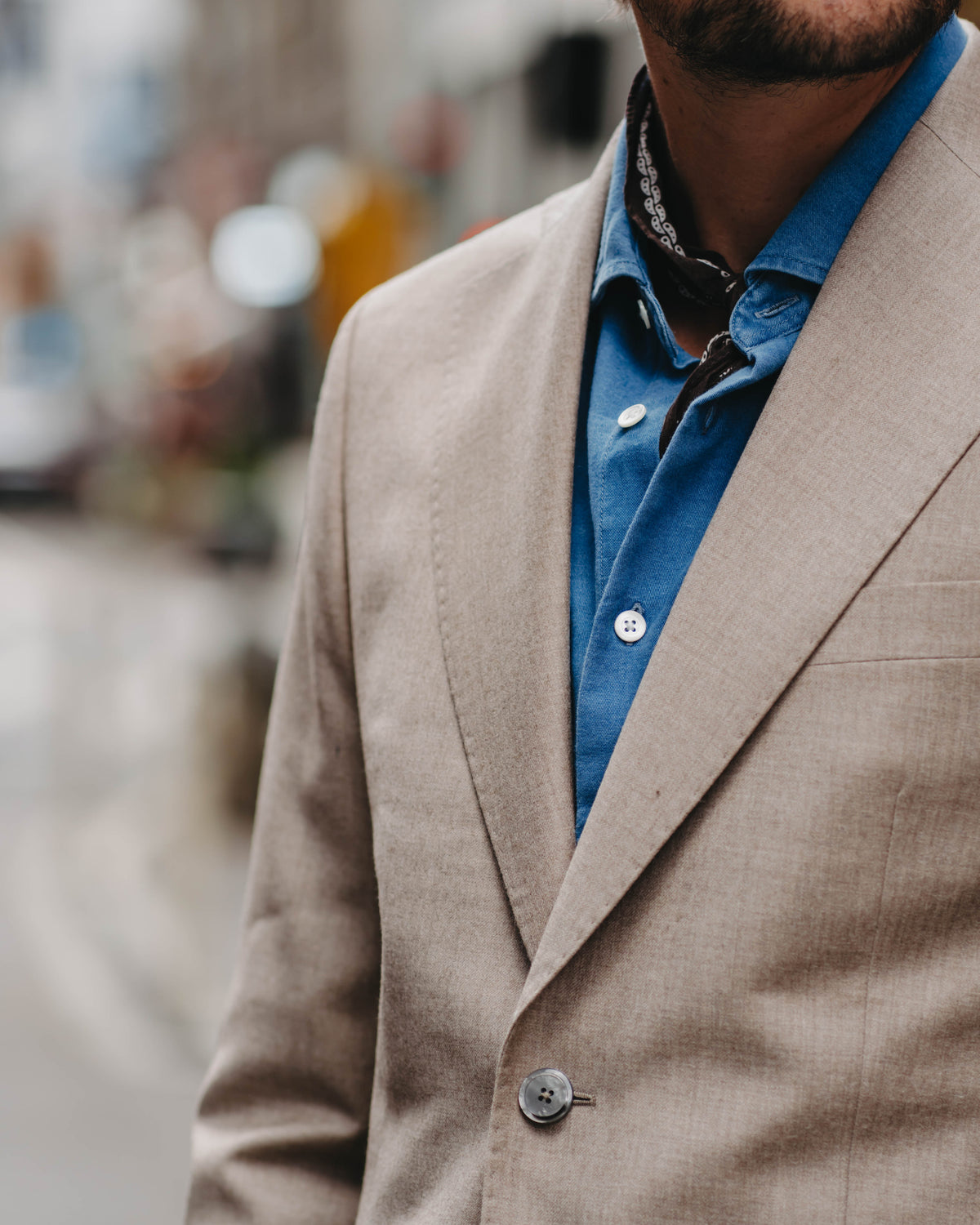 A close-up lifestyle shot of a man wearing a light beige suit jacket over a blue shirt, with a blurred outdoor street background.