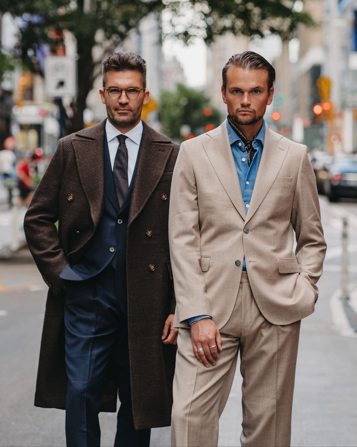 Two men walking side-by-side on a city street, showcasing formal menswear. The man on the left wears a dark brown double-breasted overcoat over a navy blue suit.