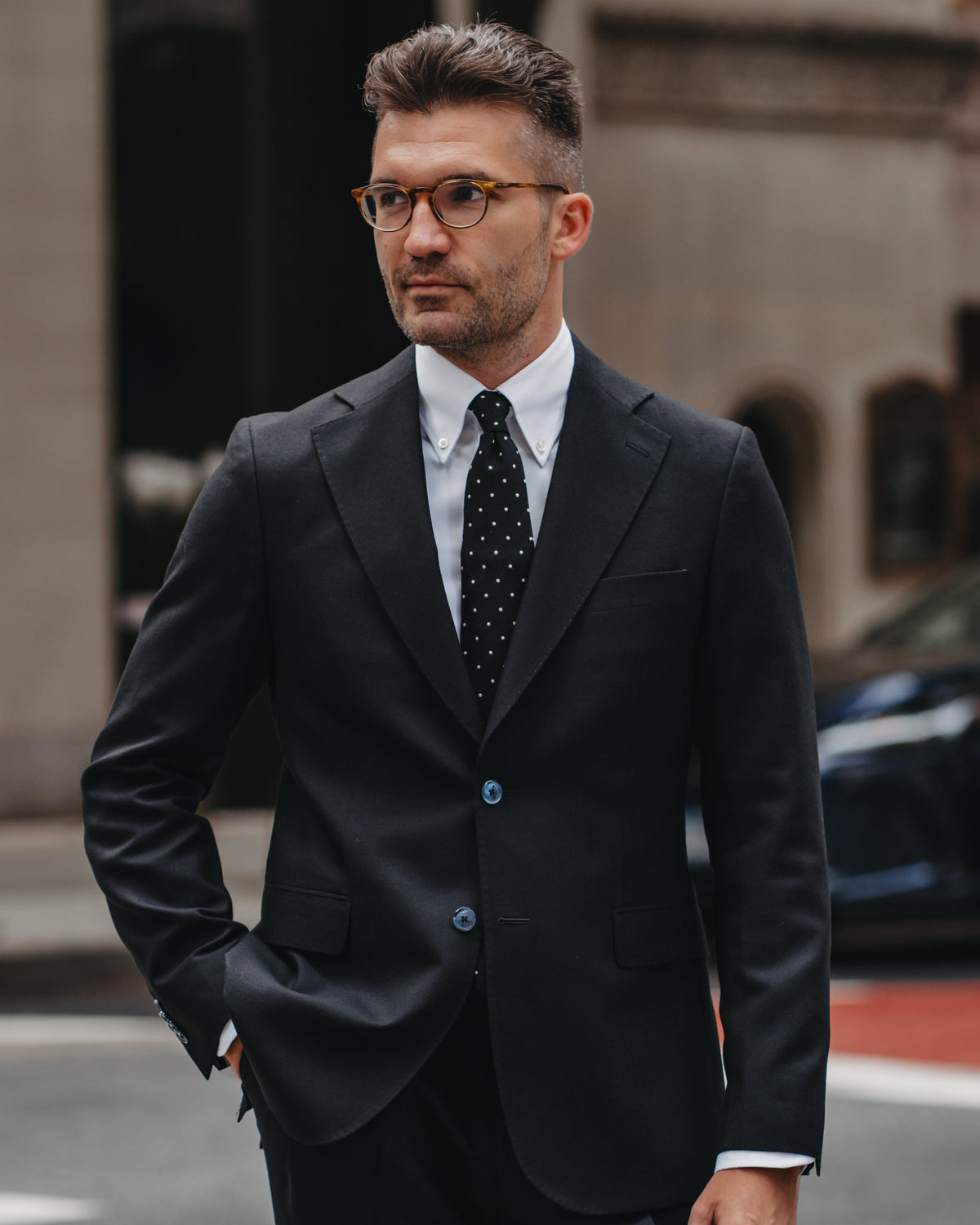 A man wearing a black two-piece suit, white dress shirt, and a black polka-dot tie, standing on a city street.
