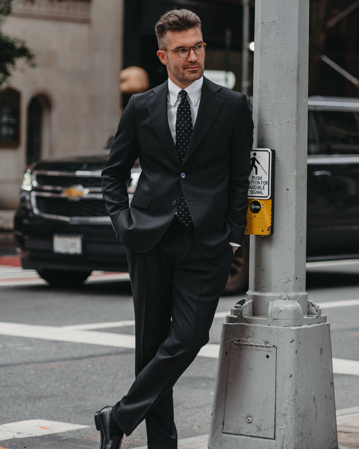 A man in a black suit, white dress shirt, and a black polka dot tie stands on a city street next to a gray pole.