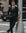 A man in a black suit, white dress shirt, and a black polka dot tie stands on a city street next to a gray pole.