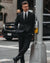 A man in a black suit, white dress shirt, and a black polka dot tie stands on a city street next to a gray pole.