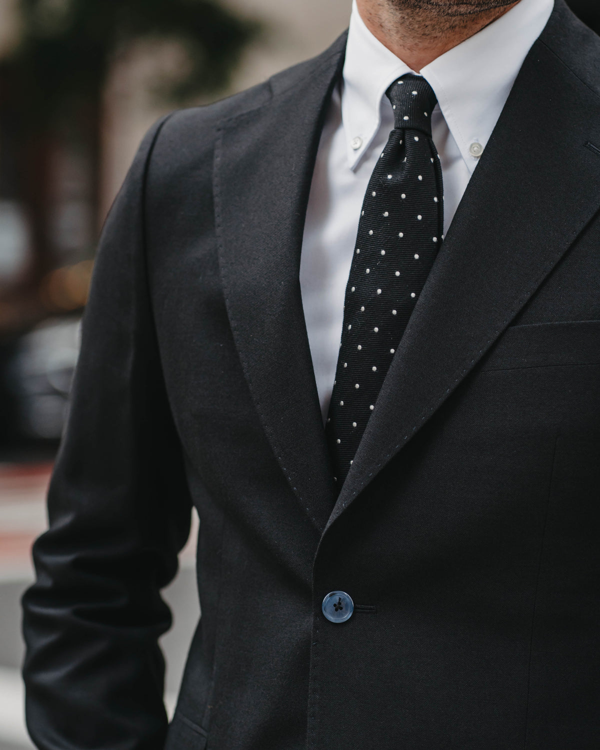 A close-up of a man wearing a black suit jacket, a crisp white dress shirt, and a black tie with small white polka dots.