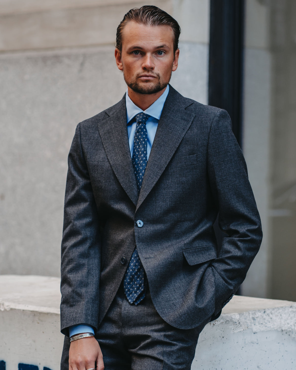 A man wearing a dark gray textured suit, a light blue dress shirt, and a blue patterned tie, standing outdoors against a light gray stone wall.