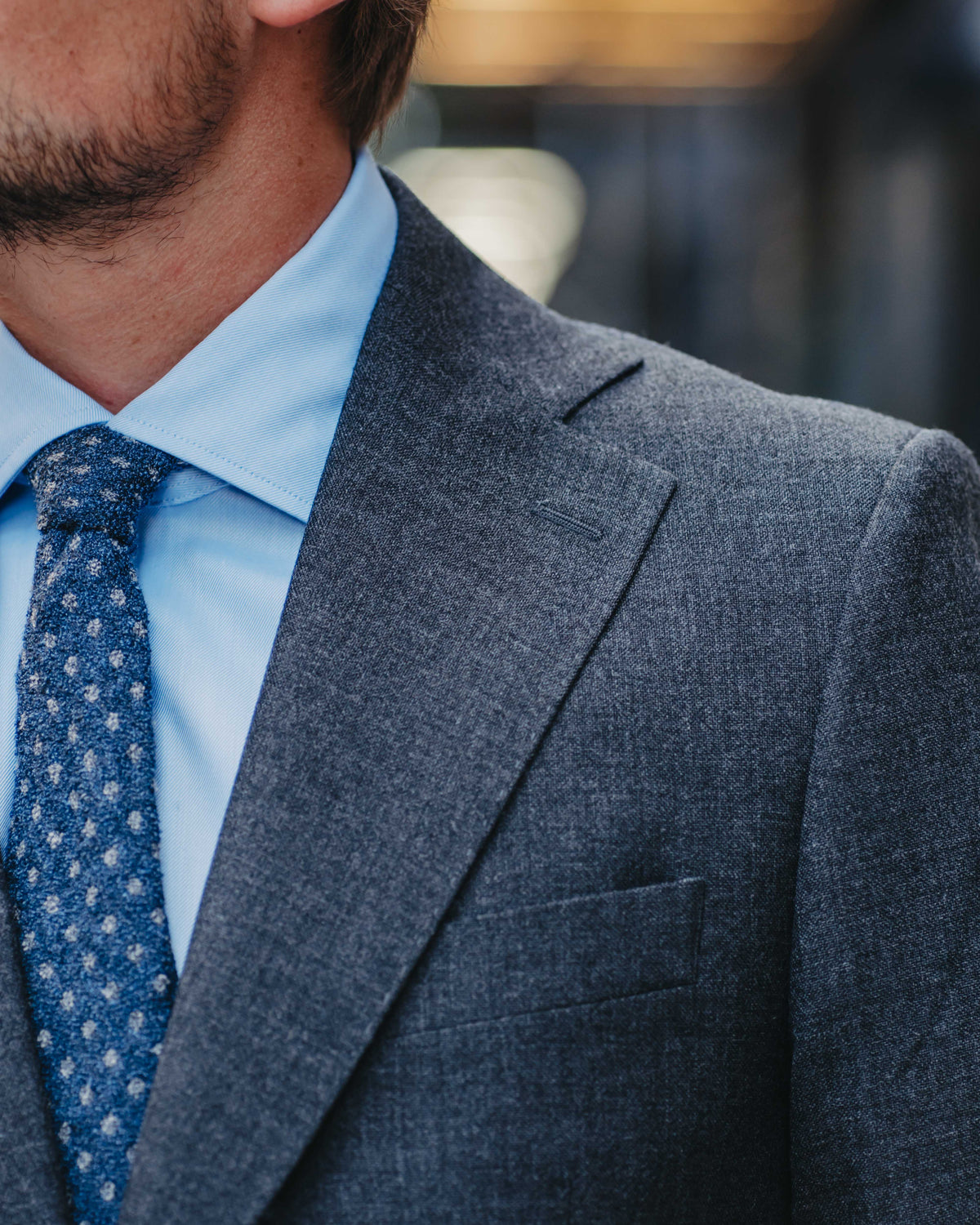 Close-up of a man wearing a textured gray suit jacket, a light blue dress shirt, and a blue patterned tie.