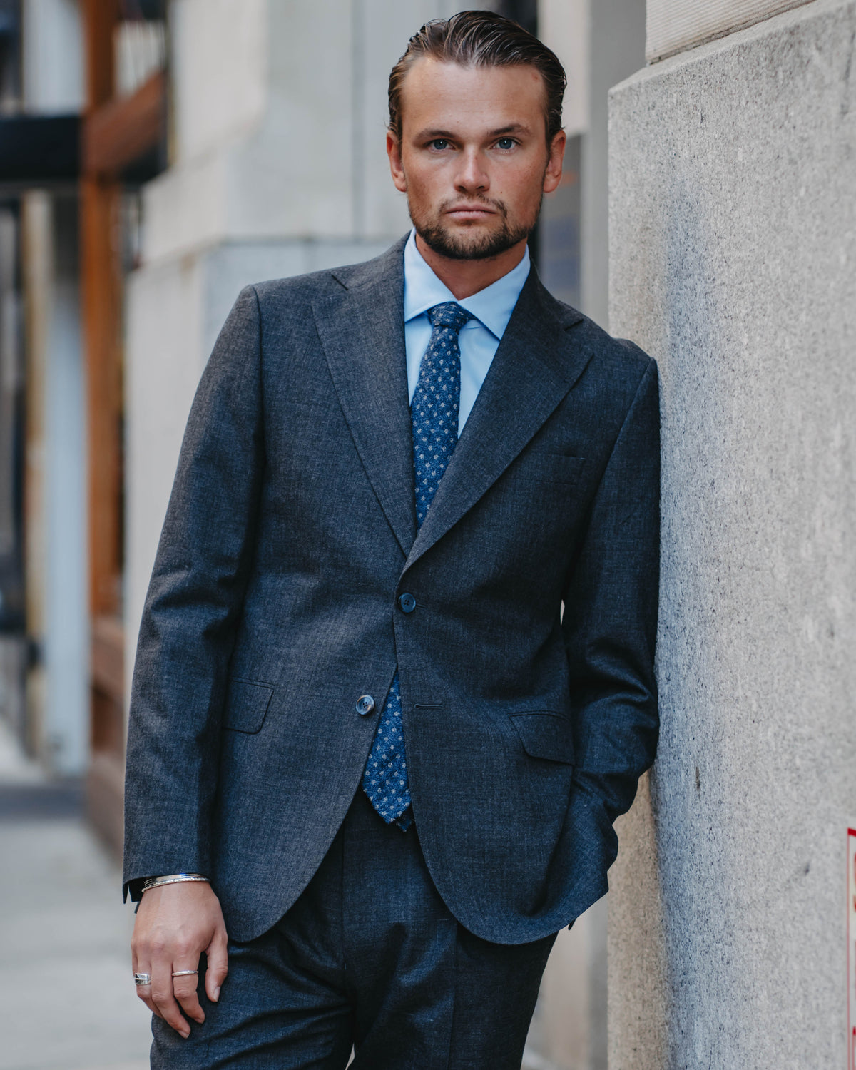 A man wearing a dark gray textured suit, a light blue dress shirt, and a blue patterned tie, standing against a stone wall in an urban setting.