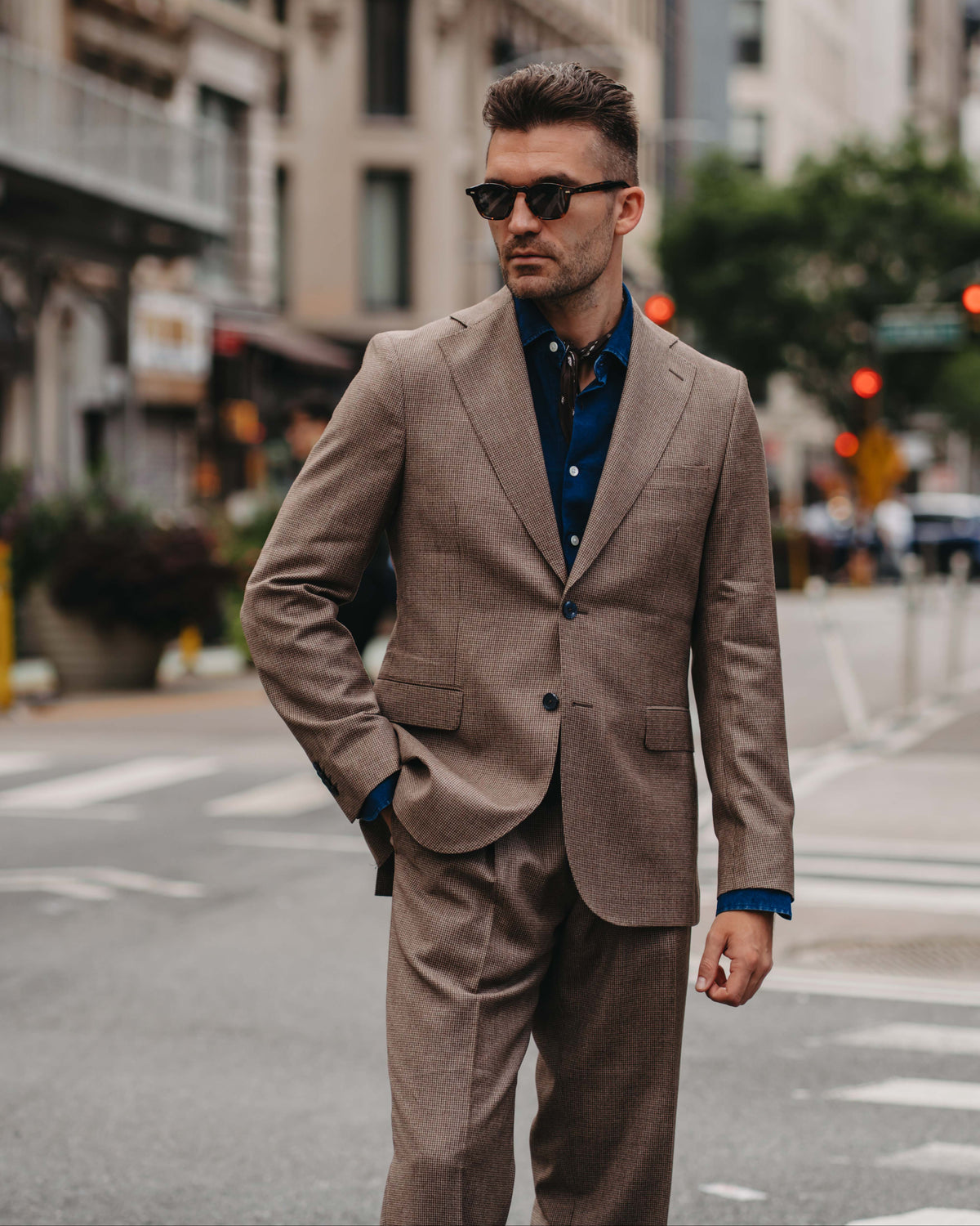 A man wearing a tailored taupe-brown suit with a dark blue button-down shirt and sunglasses, standing on a city street.