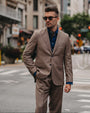 A man wearing a tailored taupe-brown suit with a dark blue button-down shirt and sunglasses, standing on a city street.