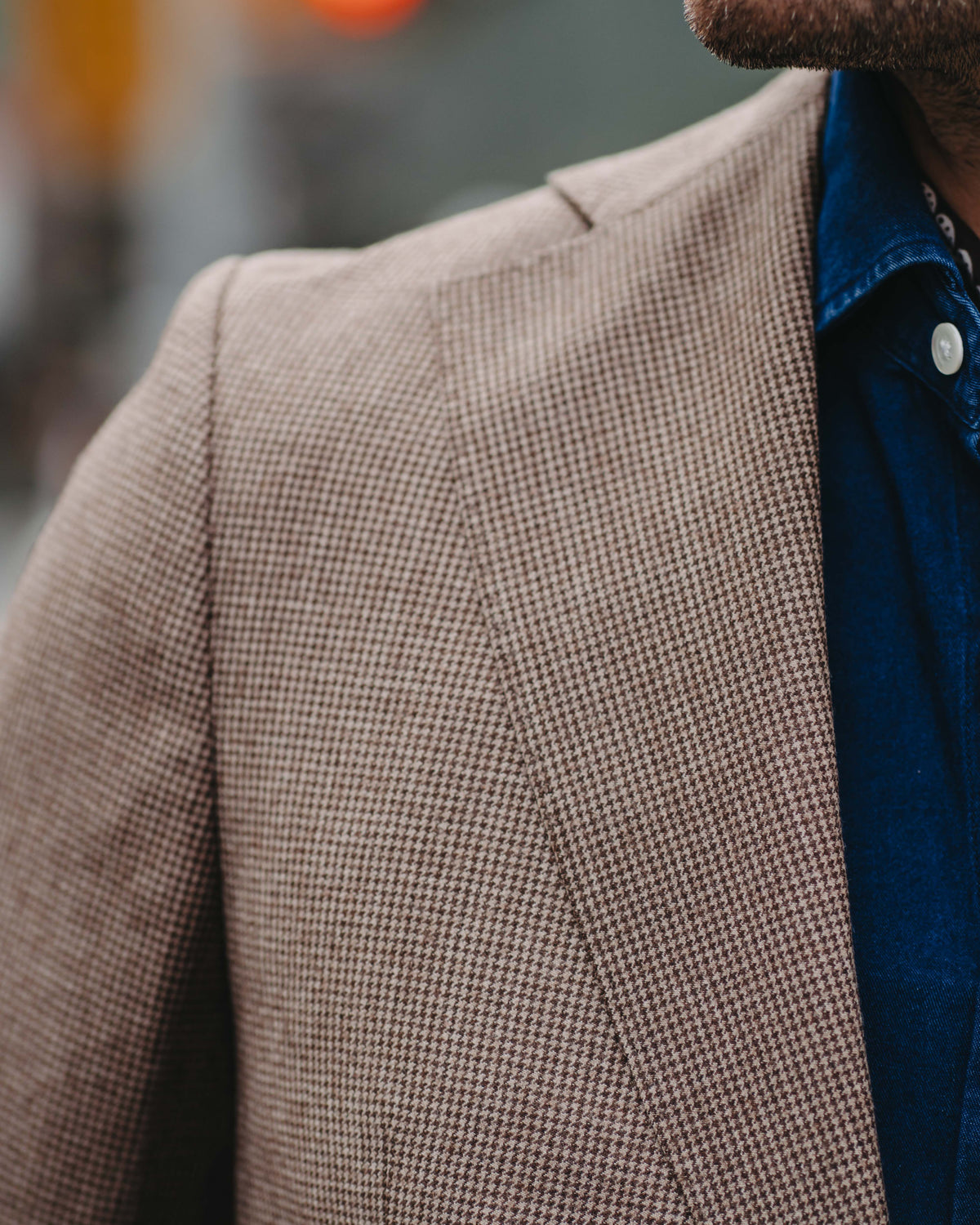 Close-up of a man wearing a beige and brown micro-check patterned blazer over a blue button-down shirt, with a blurred city street background.