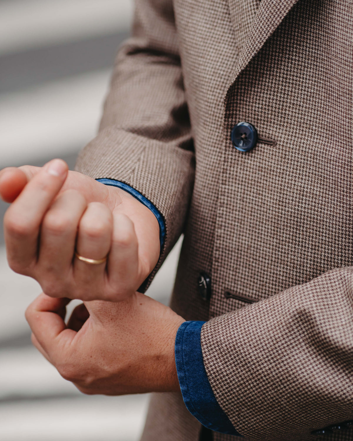 Close-up of a person wearing a brown textured blazer with a subtle houndstooth pattern, a blue shirt cuff, and a gold ring on one finger.