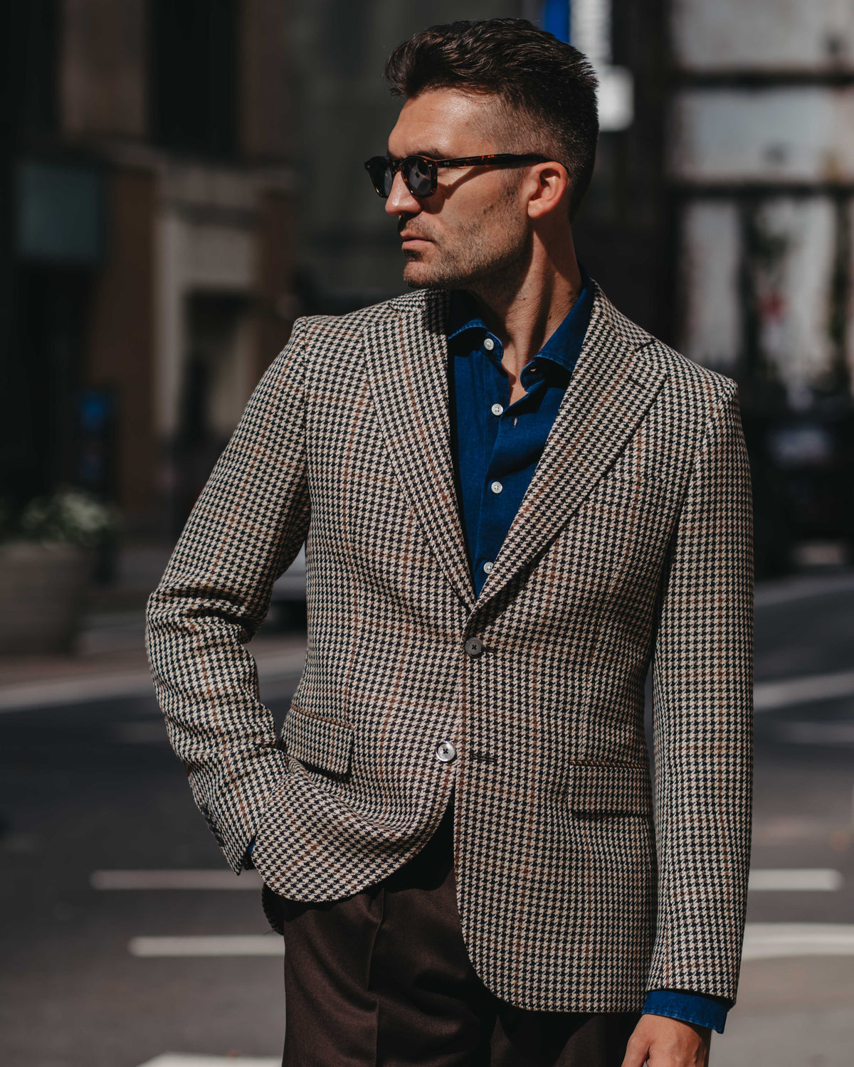 A man wearing a brown and beige houndstooth blazer over a blue button-down shirt, styled with dark brown trousers and sunglasses, standing on an urban street.