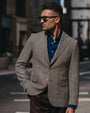 A man wearing a brown and beige houndstooth blazer over a blue button-down shirt, styled with dark brown trousers and sunglasses, standing on an urban street.