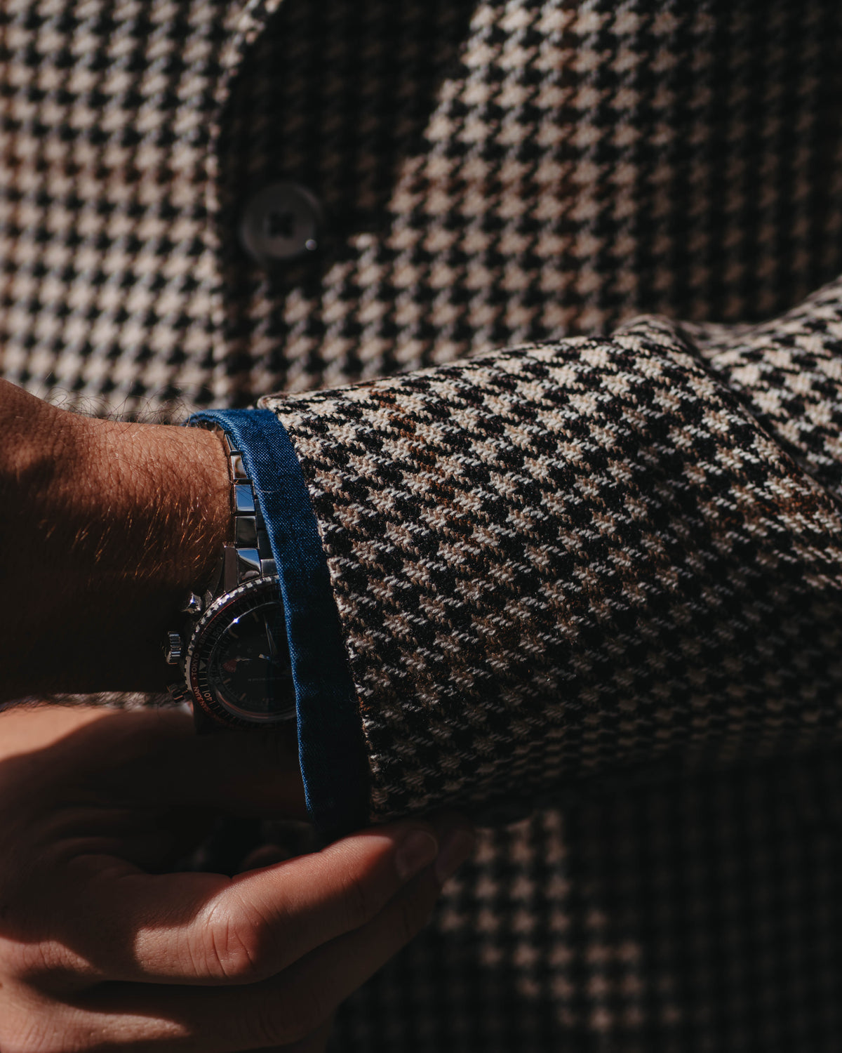 A close-up lifestyle shot of a person wearing a black and beige houndstooth blazer, adjusting the cuff of the sleeve to reveal a bright blue denim watch strap and a dark metal watch.