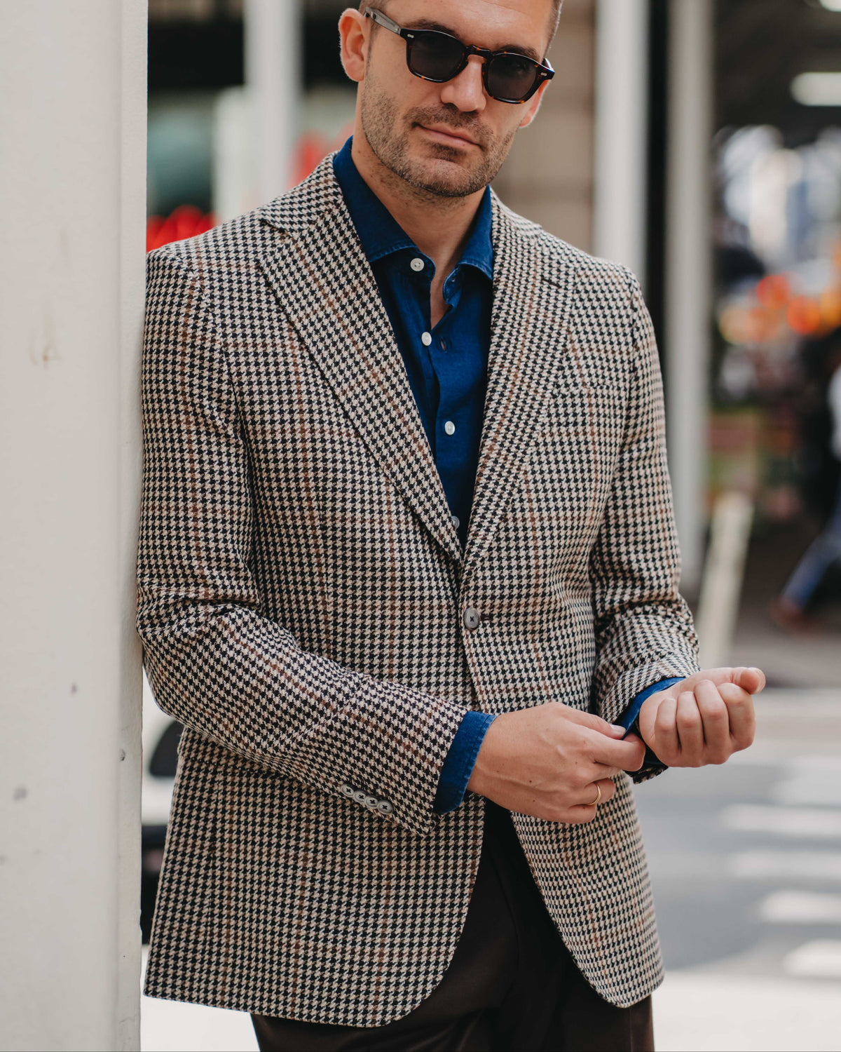 A man wearing a brown and beige houndstooth blazer over a dark blue button-down shirt, styled with black sunglasses and dark trousers, standing in an urban outdoor setting.