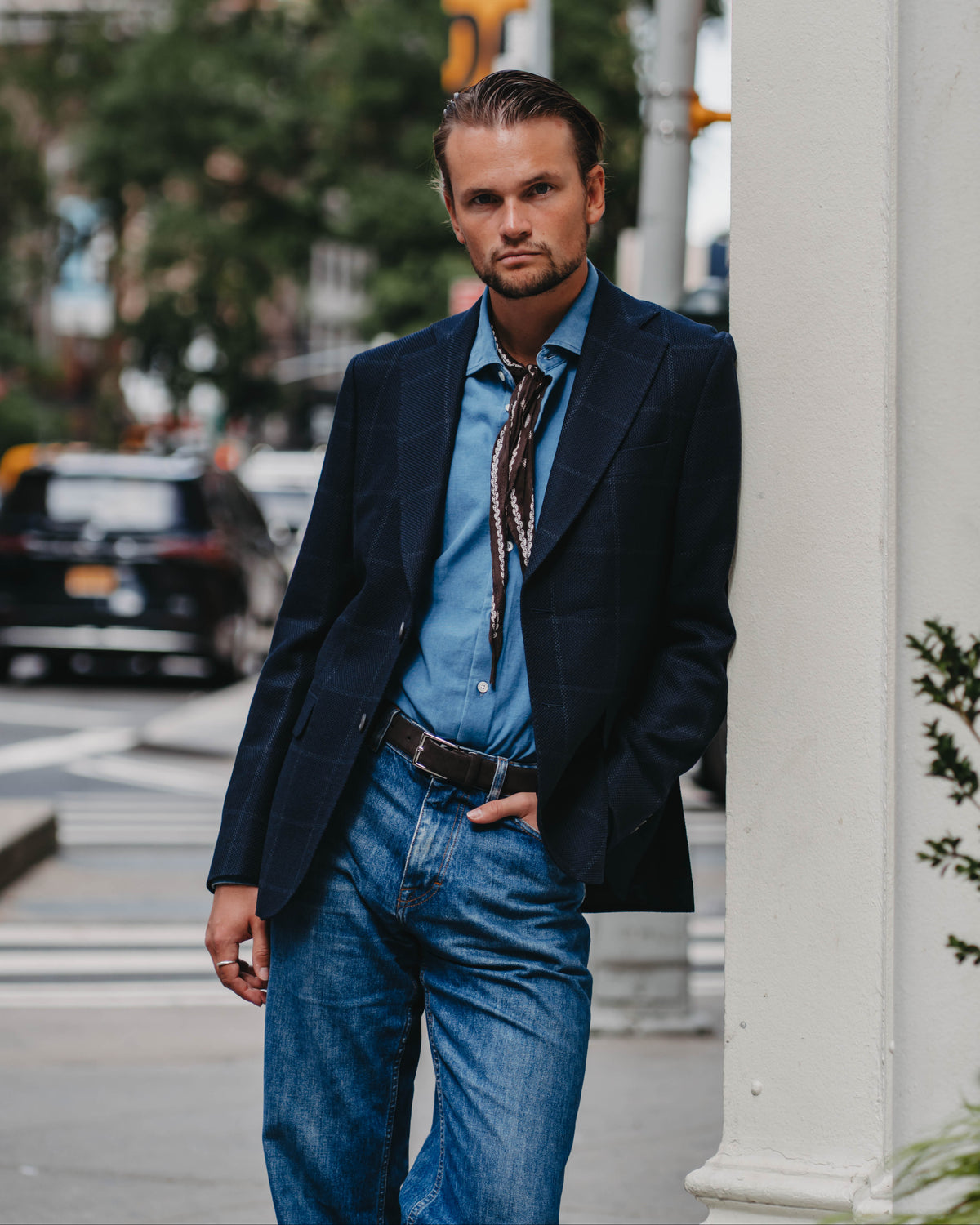 A man wearing a dark navy plaid blazer, a light blue button-down shirt, a brown patterned necktie, and blue jeans, standing on a city street next to a white column.