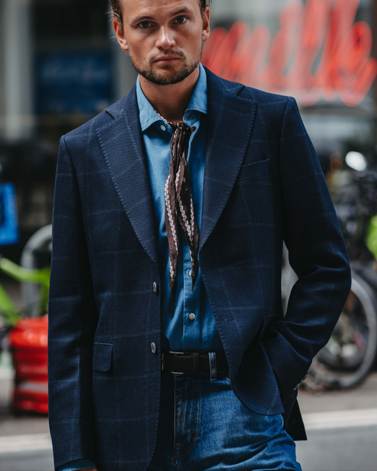 A man wearing a dark blue plaid blazer, a blue button-down shirt, and a patterned brown necktie, standing in front of a blurred city street background.