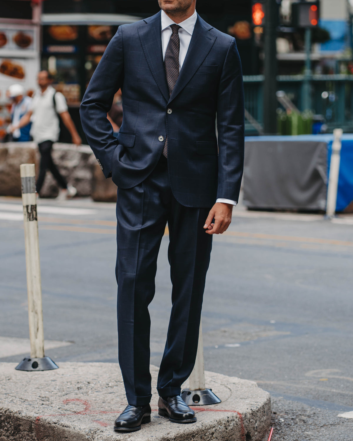 A man wearing a navy blue suit, white dress shirt, dark tie, and sunglasses stands on a city sidewalk corner with his hands in his pockets, with a blurred urban street background.