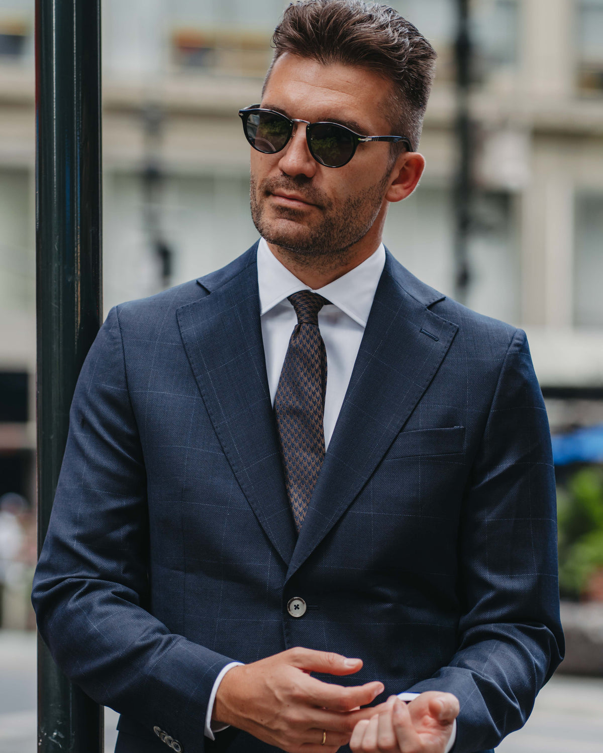 A man wearing a navy blue windowpane suit, white dress shirt, patterned tie, and dark sunglasses, standing outdoors next to a black pole.