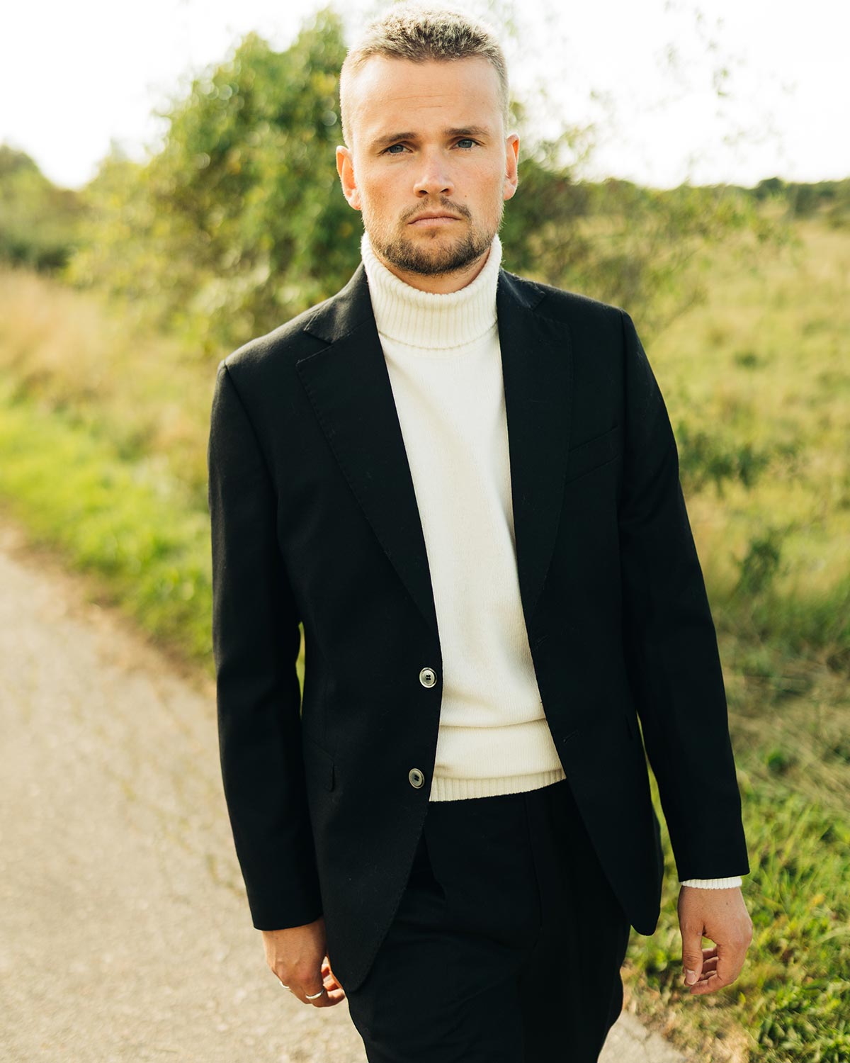 Man wearing a black blazer over a white turtleneck sweater in a natural setting.