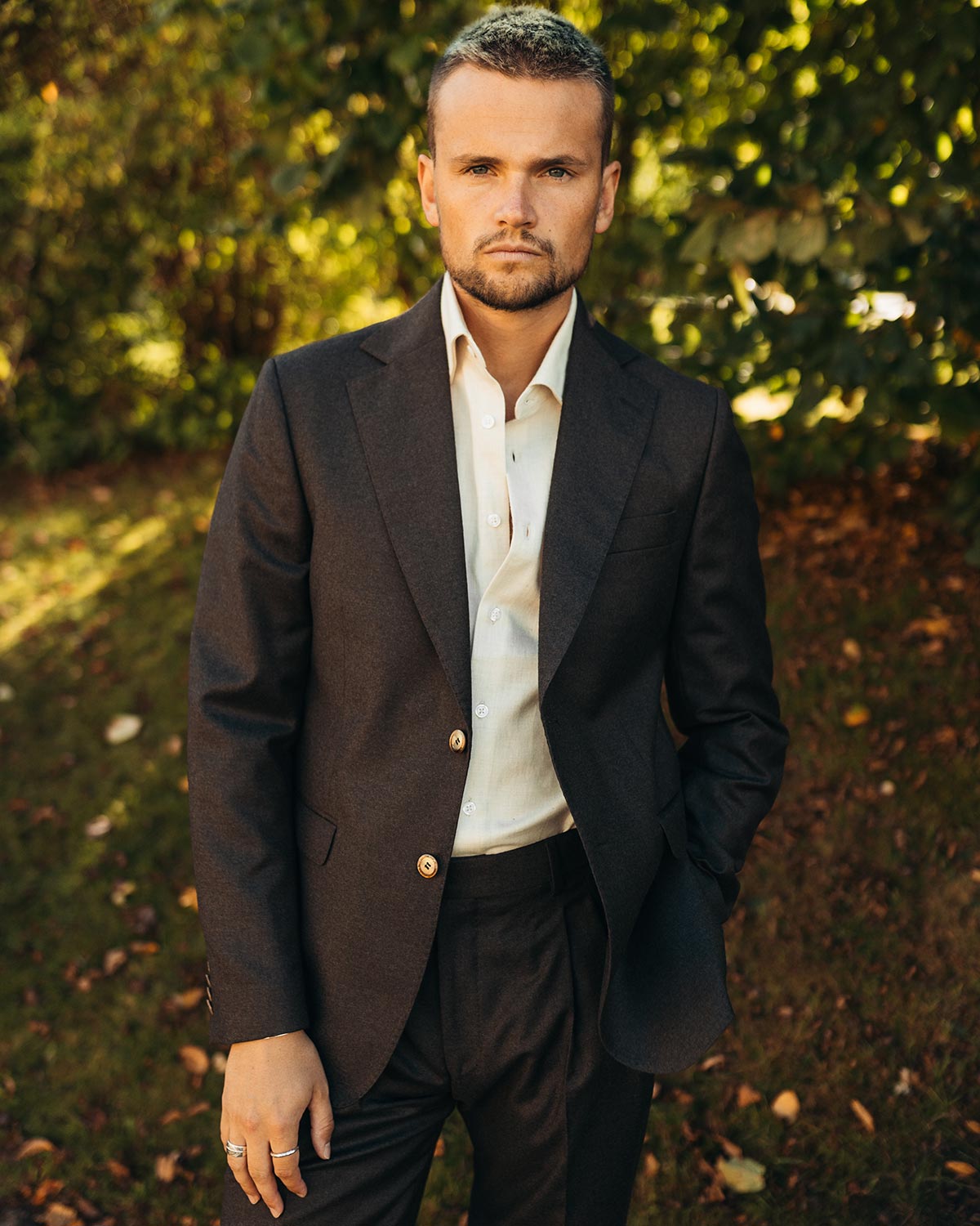 Man in a brown flannel suit standing outdoors with greenery in the background
