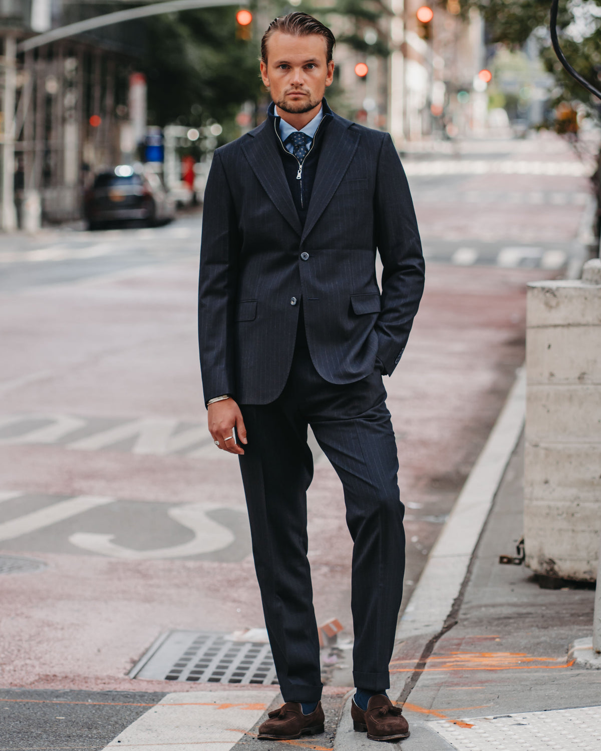 A man wearing a dark navy blue pinstripe suit, light blue dress shirt, and patterned tie stands on a city street crosswalk.