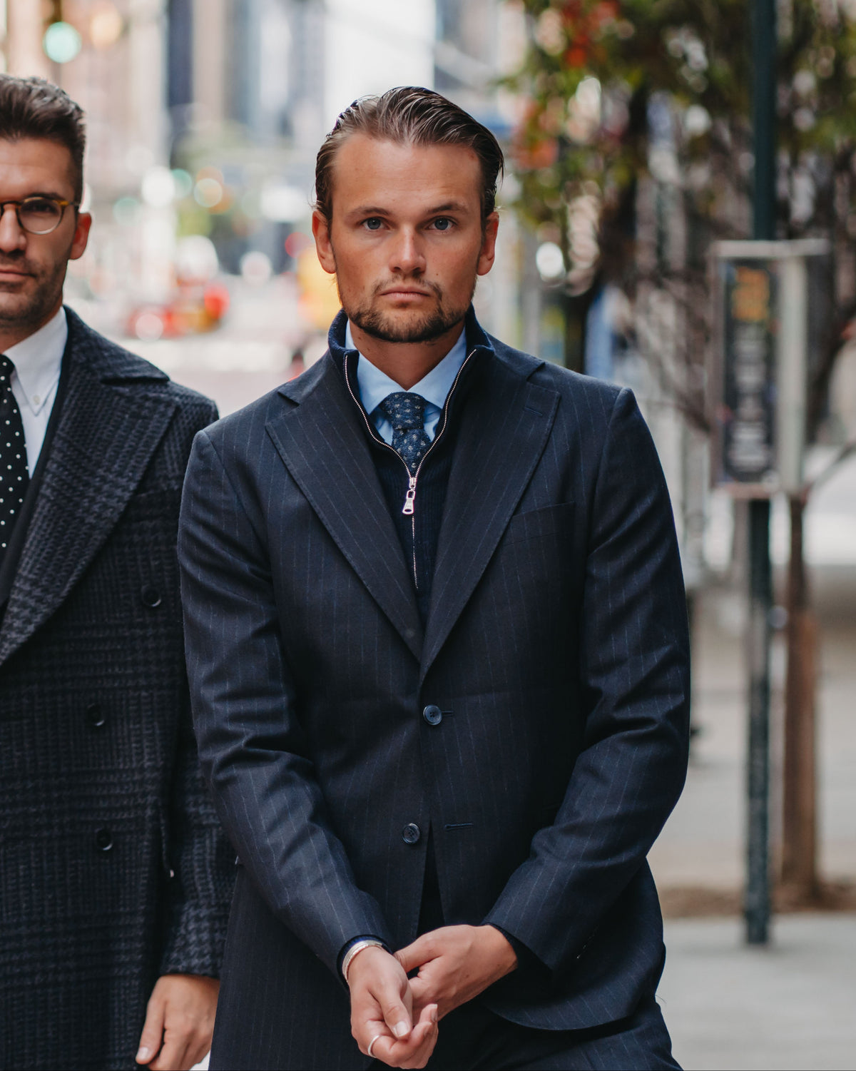 Two men in tailored suits standing on a city street. The man on the left wears a dark charcoal plaid double-breasted overcoat with a white shirt and black patterned tie.