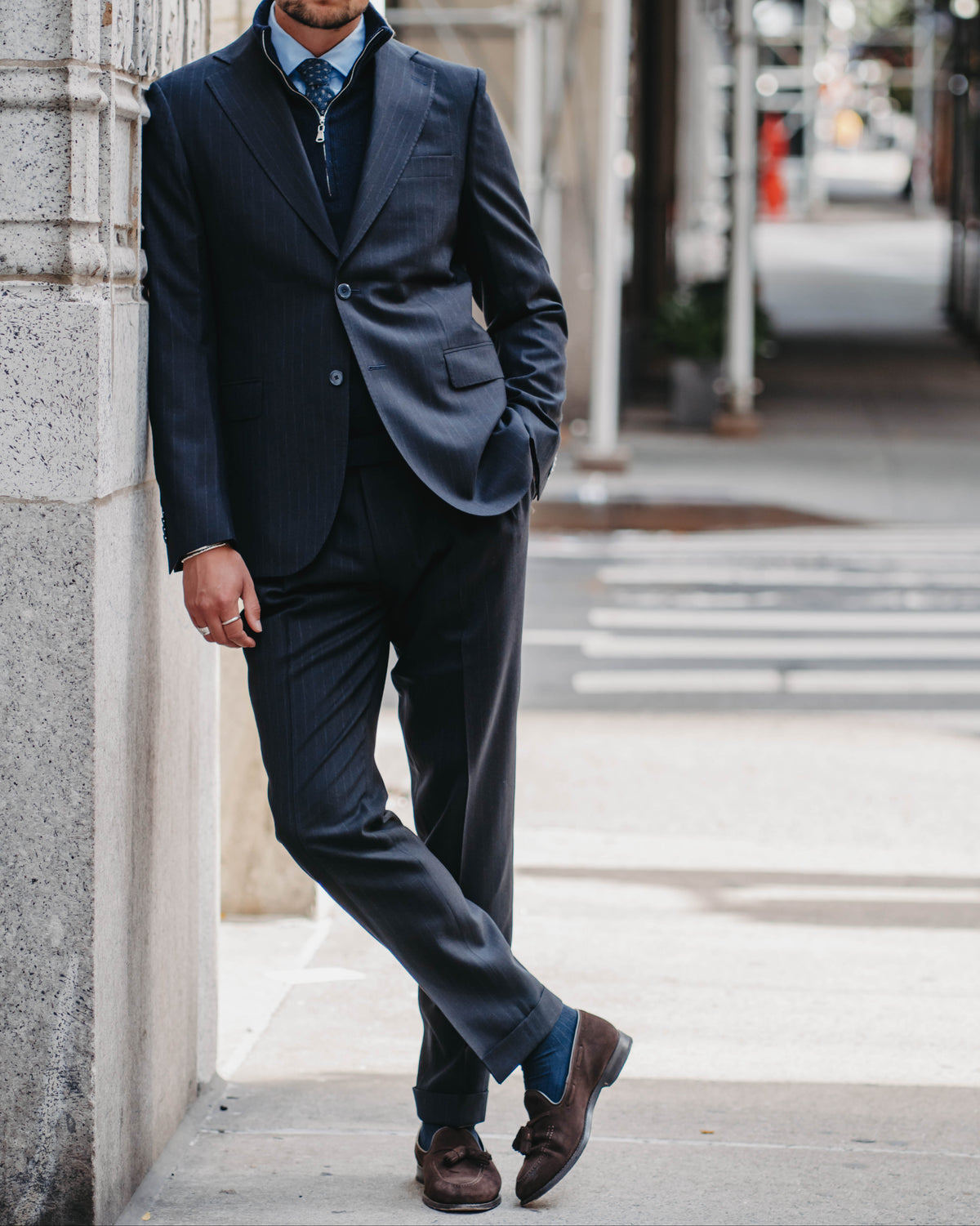 A man wearing a dark navy pinstripe suit, light blue dress shirt, and patterned tie leans against a stone building on a city sidewalk.