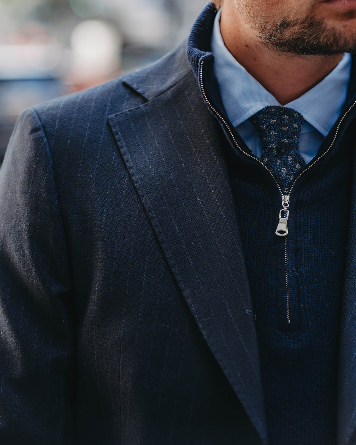 A close-up shot of a man wearing a dark navy blue pinstripe suit jacket over a matching zip-up sweater, layered over a light blue dress shirt and a patterned tie.