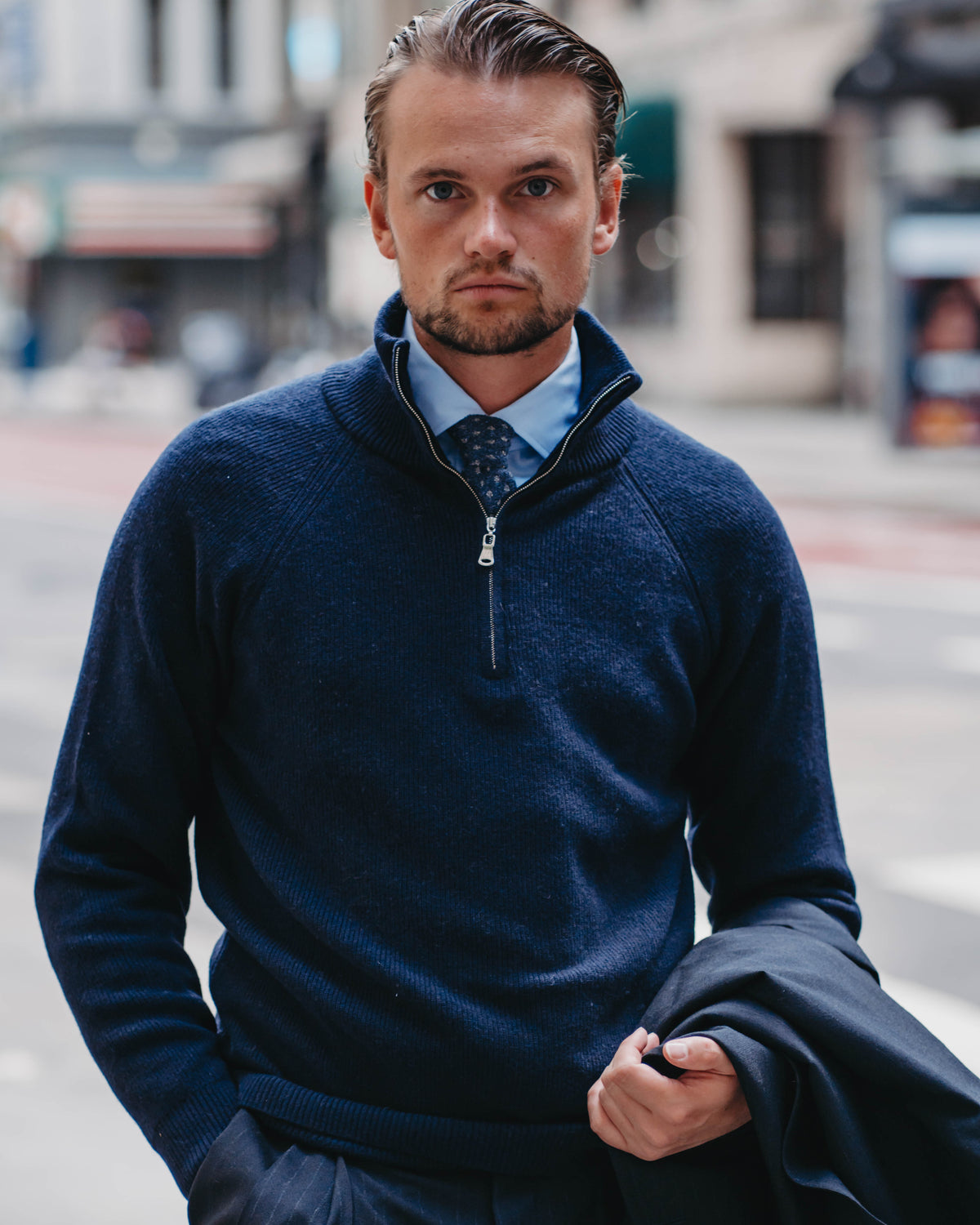 A man wearing a navy blue quarter-zip sweater over a light blue dress shirt and patterned tie, standing on a city street and holding a dark jacket.