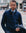 A man wearing a navy blue quarter-zip sweater over a light blue dress shirt and patterned tie, standing on a city street and holding a dark jacket.