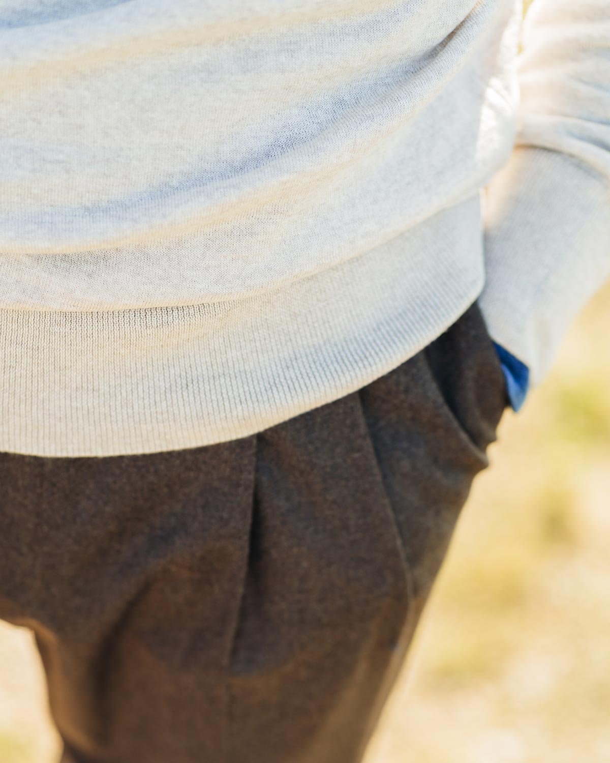 Close-up of a person wearing a light beigesweater and brown pants with a blurred natural background.