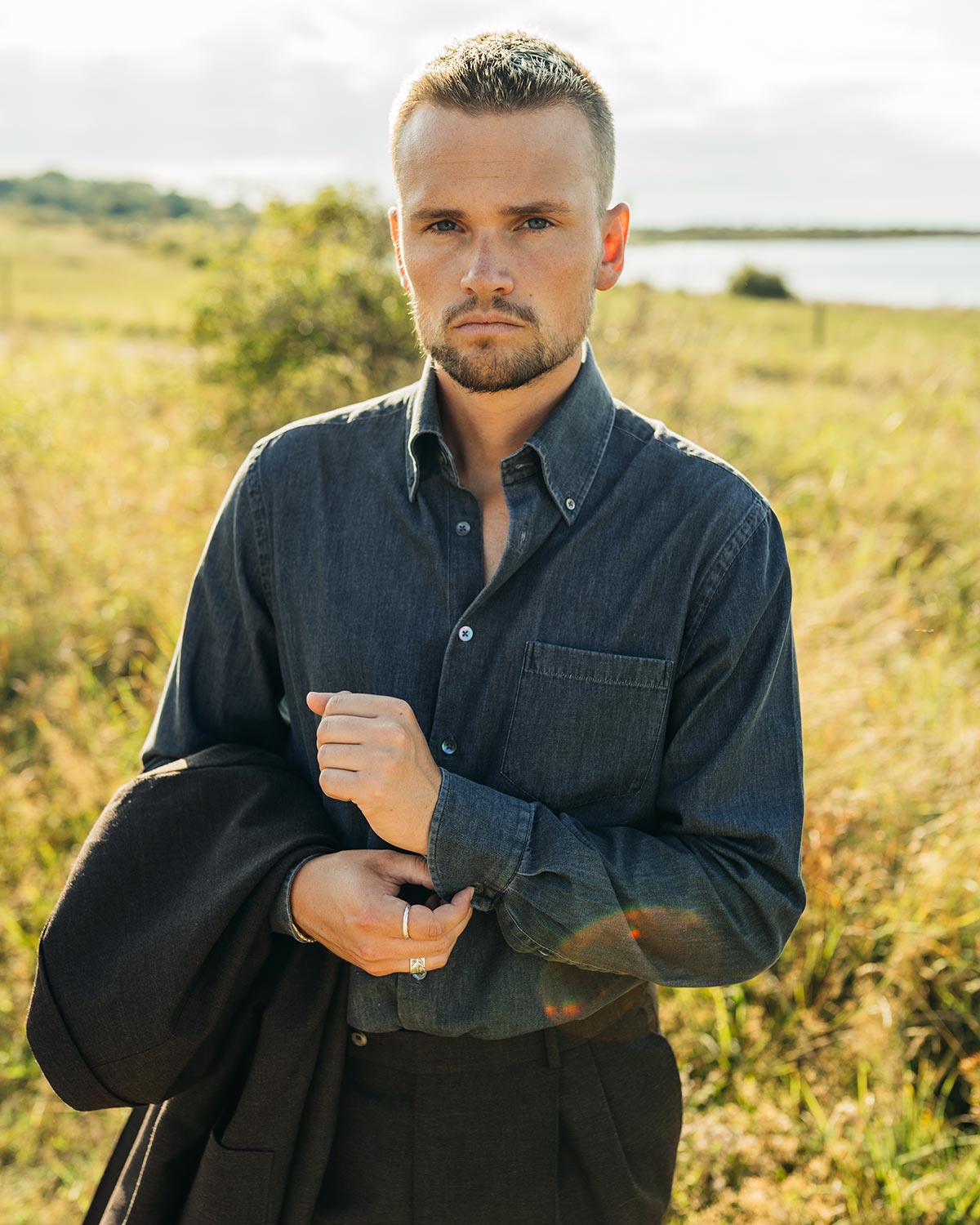 Man standing in a field wearing a dark denim shirt and holding a jacket.