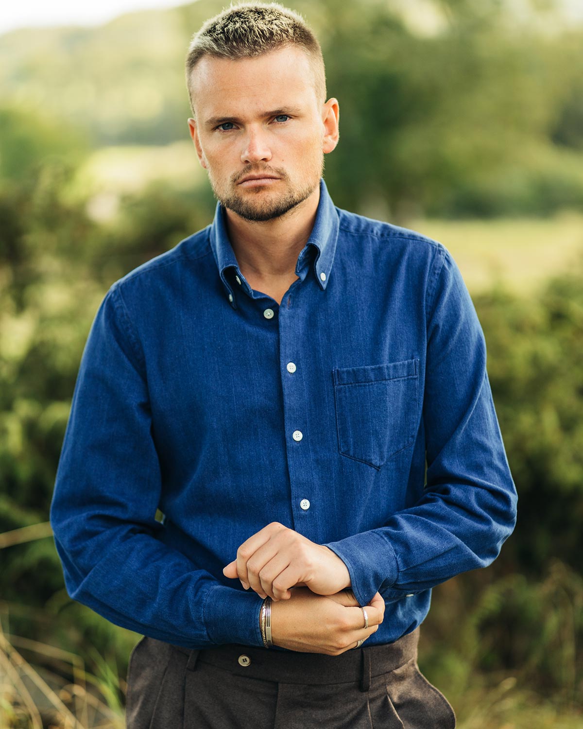 Man wearing a blue denim shirt standing outdoors with greenery in the background