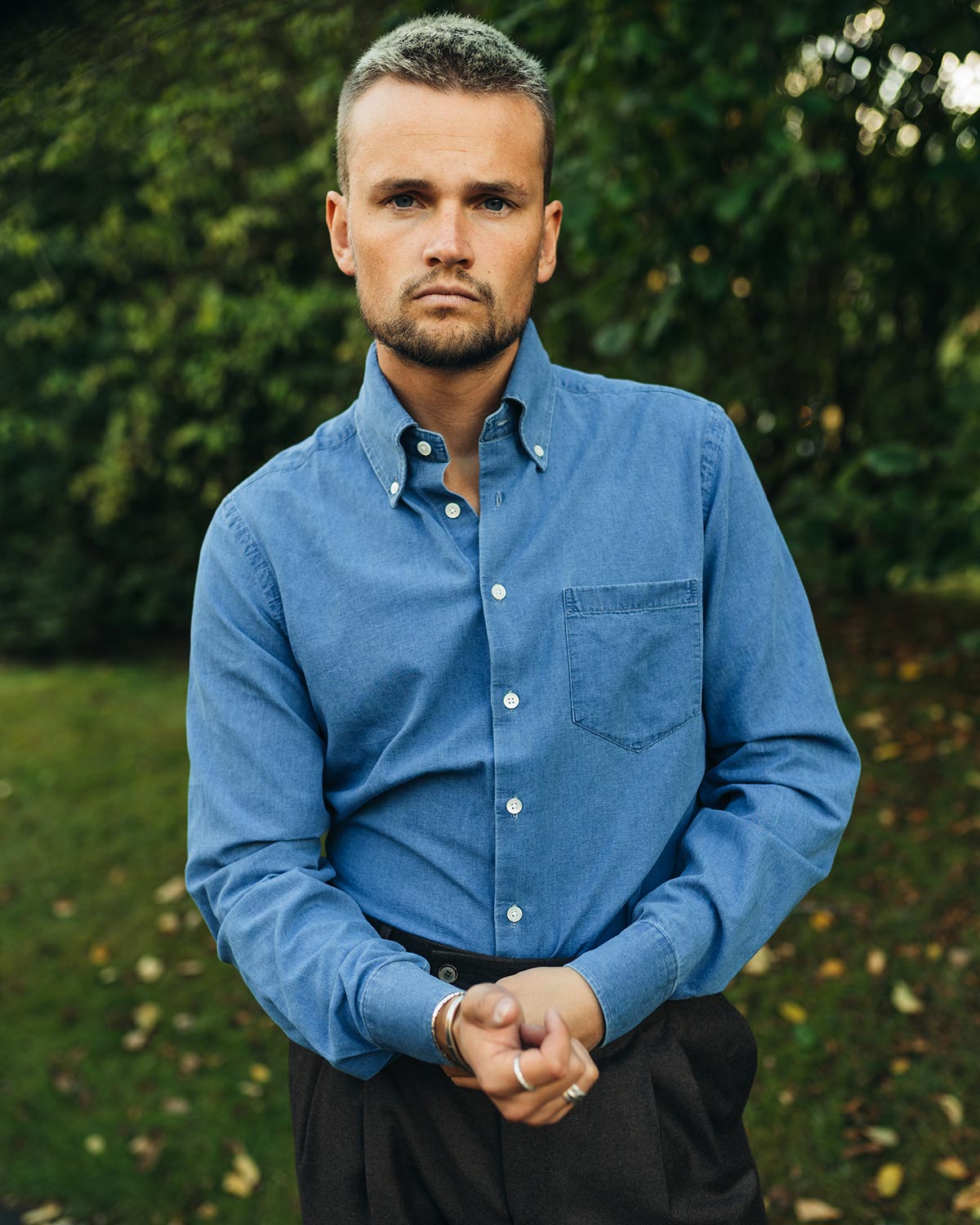Man wearing a blue denim shirt standing outdoors with greenery in the background